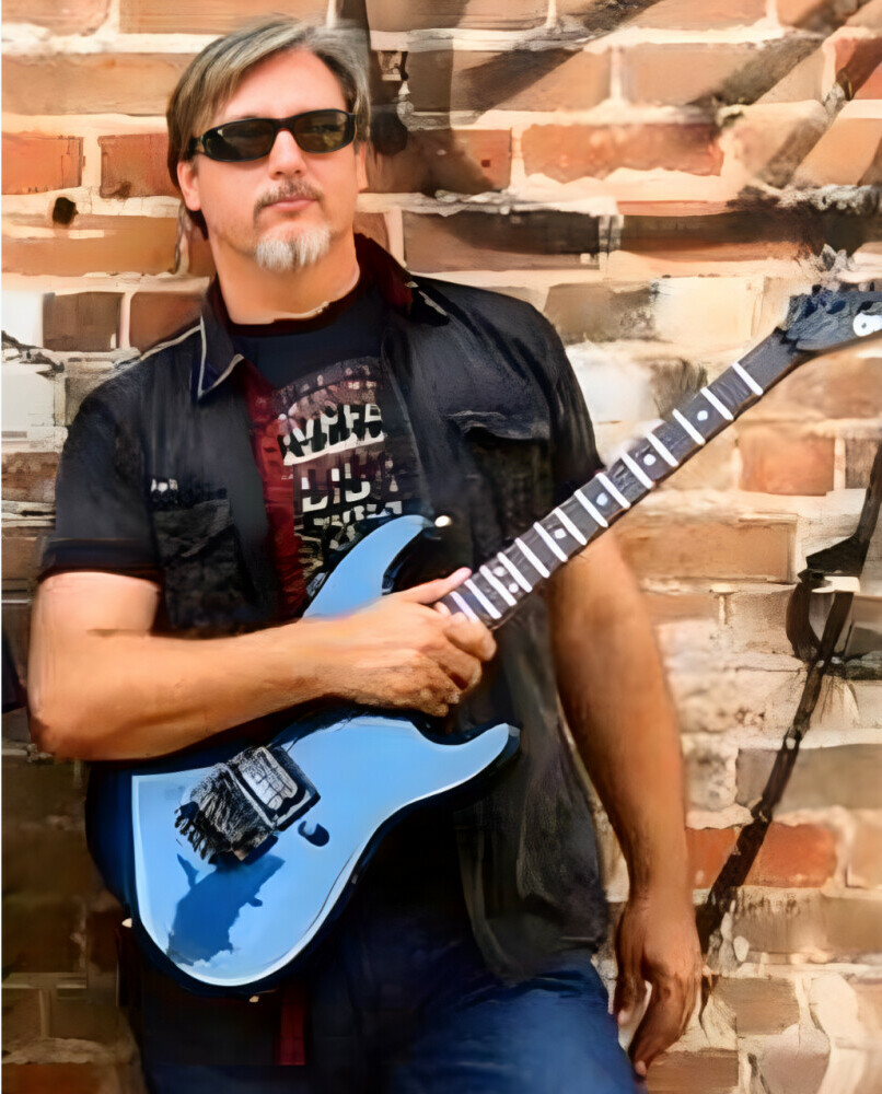 Guy leaning against a brick wall holding a blue 1987 Charvel electric guitar
