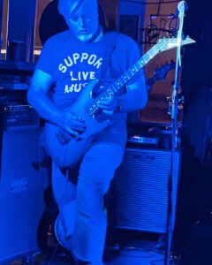 Playing a blue charvel guitar wearing a Support Live Music t-shirt