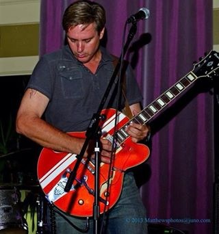 Guy playing an orange color semi-hollow Ibanez with white stripes live on stage. 